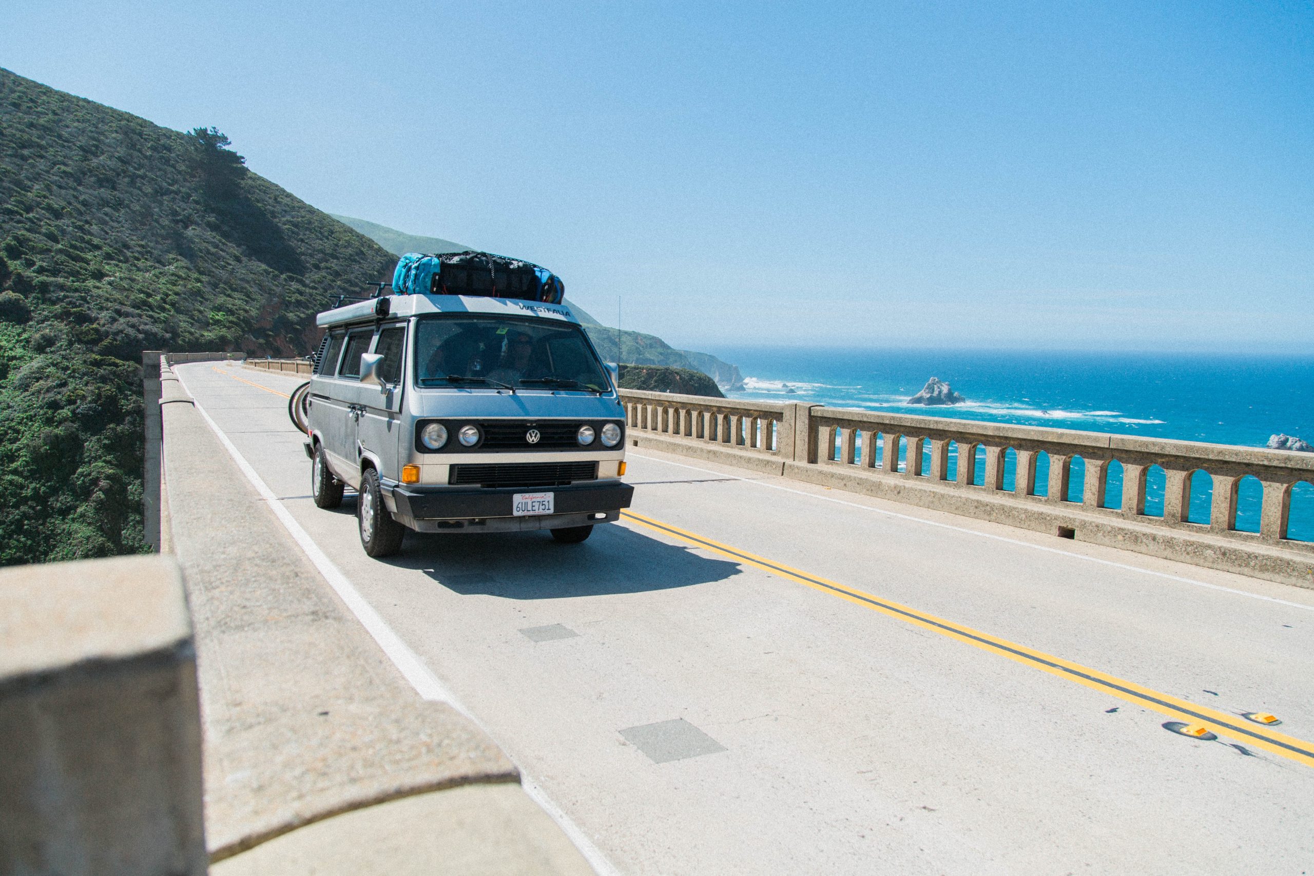 dark blue VW camper van driving along beach road, with blue ocean in the background, loaded up for a drip with bikes on the back
