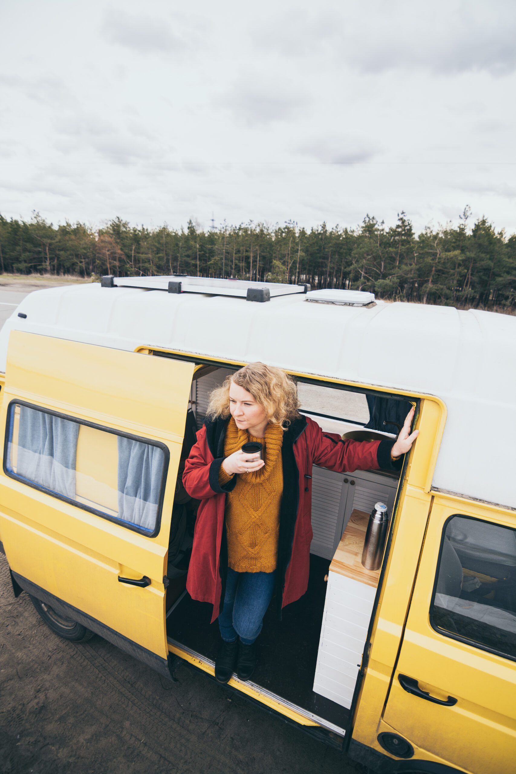 Young blond woman traveling by camper van with solar panel on the roof top and pine forest on the background.